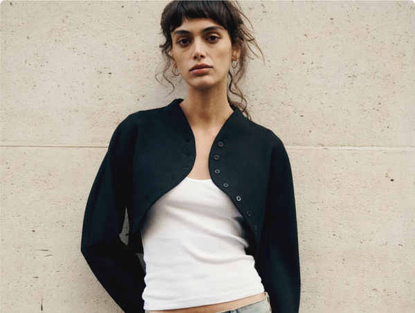A woman with styling a structured, curved black bolero jacket featuring delicate button details over a classic white tank top.
