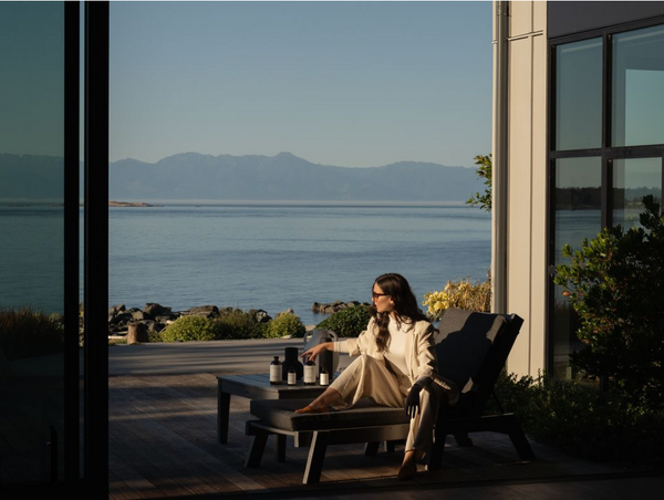 A woman in a cream-colored pantsuit relaxing on an outdoor patio lounge chair overlooking a calm ocean and distant mountains.