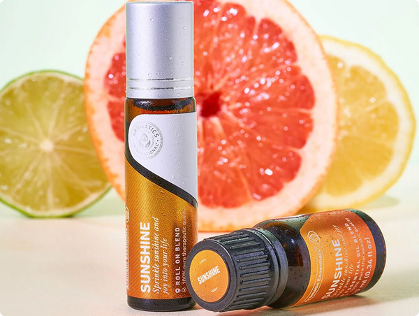 Aromatics International Sunshine roll-on and essential oil bottles displayed in front of sliced fresh grapefruit, lime, and lemon.