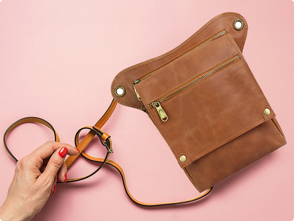 A hand with vibrant red nails holding the strap of a brown leather utility hip bag.
