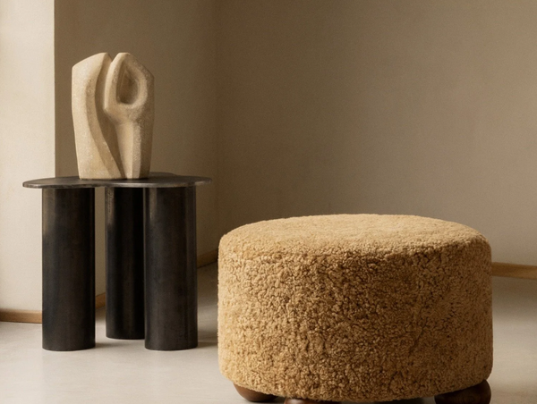 A simple room setting showing a round, textured beige pouf ottoman next to a dark metal side table with a modern stone sculpture.