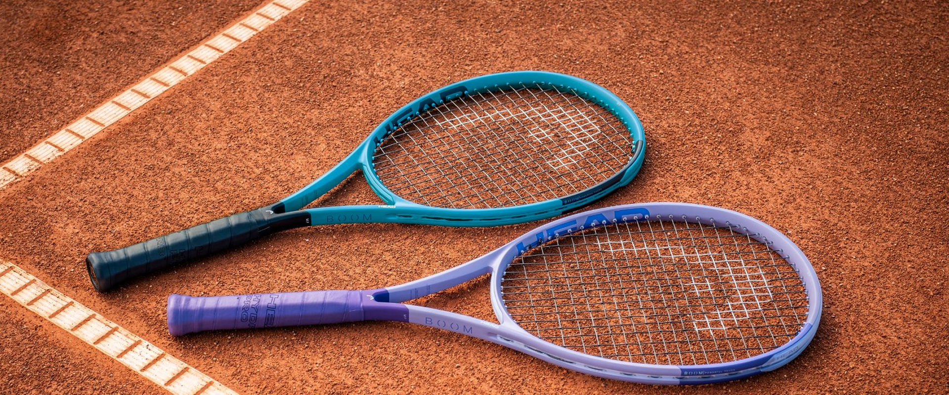 Two Head Boom tennis racquets, one mint green and one lavender purple, laying flat on a red clay court