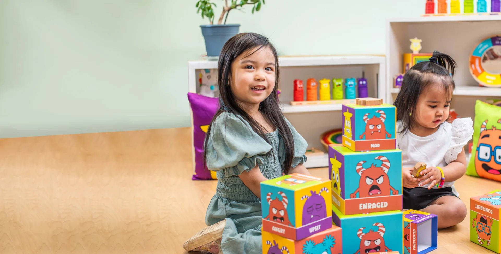 Two young girls happily playing together on the floor with colorful, stacking emotion blocks featuring expressive cartoon monsters