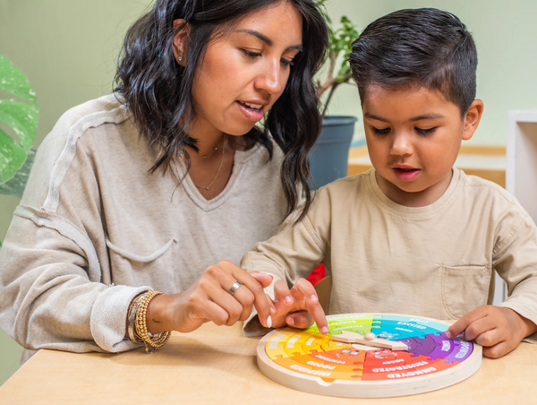 A mother gently guiding a young boy as he points to different sections of a colorful wooden emotion wheel toy at a table.