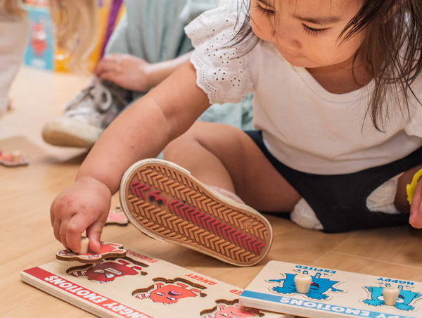 A close-up shot of a young girl focused on placing a wooden monster puzzle piece labeled "Angry" into an "Angry Emotions" board on the floor.