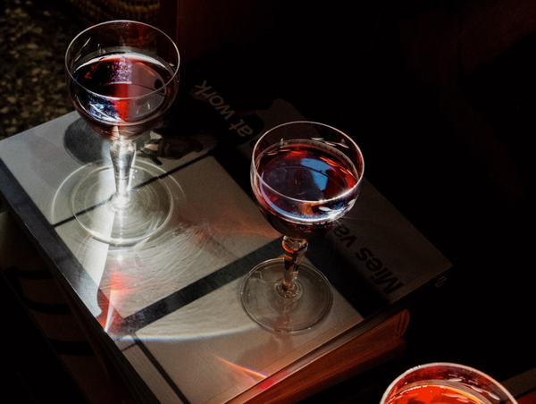 A moody, overhead lifestyle image showing two elegant crystal glasses filled with a deep red aperitif, resting on top of a book