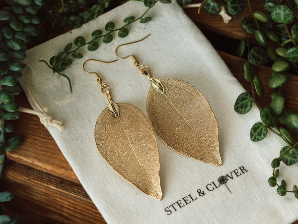 A pair of gold-toned, delicate metal leaf earrings from Steel & Clover, resting on a branded white fabric pouch surrounded by greenery.