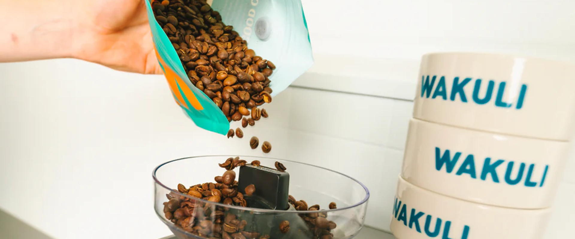A horizontal lifestyle banner image showing whole coffee beans pouring into a clear grinder hopper next to a stack of branded mugs.