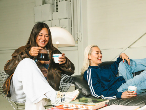 A cozy lifestyle image of two friends laughing together indoors while sharing a fresh pot of coffee in branded Wakuli cups.