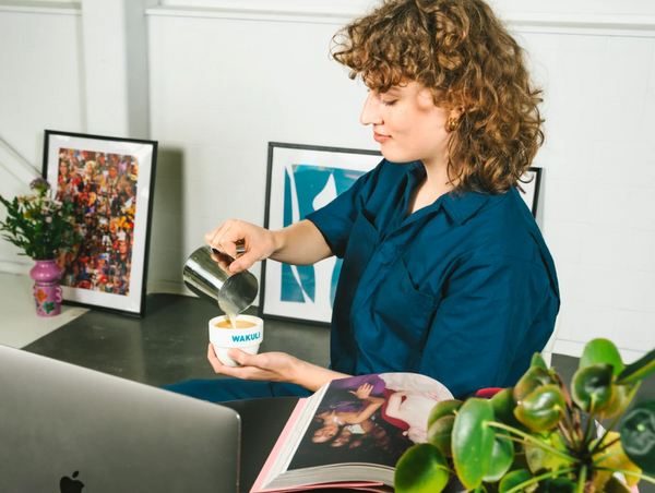A horizontal lifestyle image of a person pouring steamed milk from a steel pitcher into a branded Wakuli cup to make a latte in office.