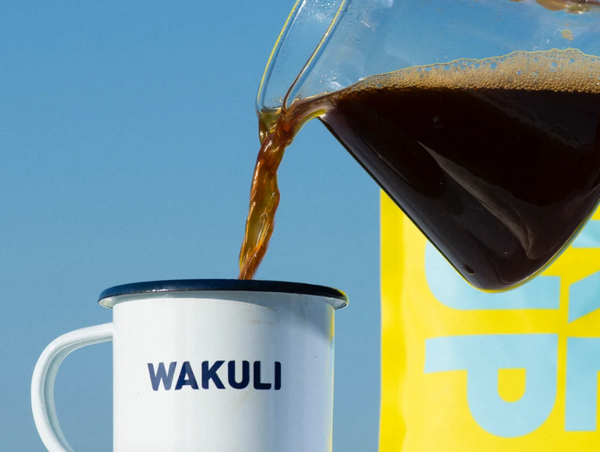 A sunny lifestyle image showing fresh coffee being poured from a glass carafe into a white enamel Wakuli camping mug.
