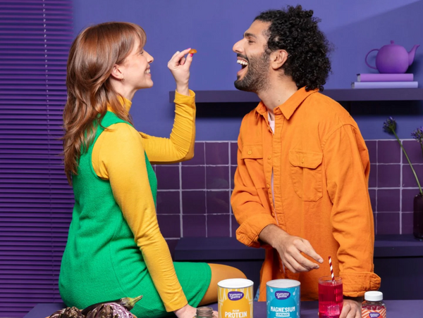 A playful lifestyle studio photograph featuring a woman sitting on a counter, feeding a gummy supplement to a man